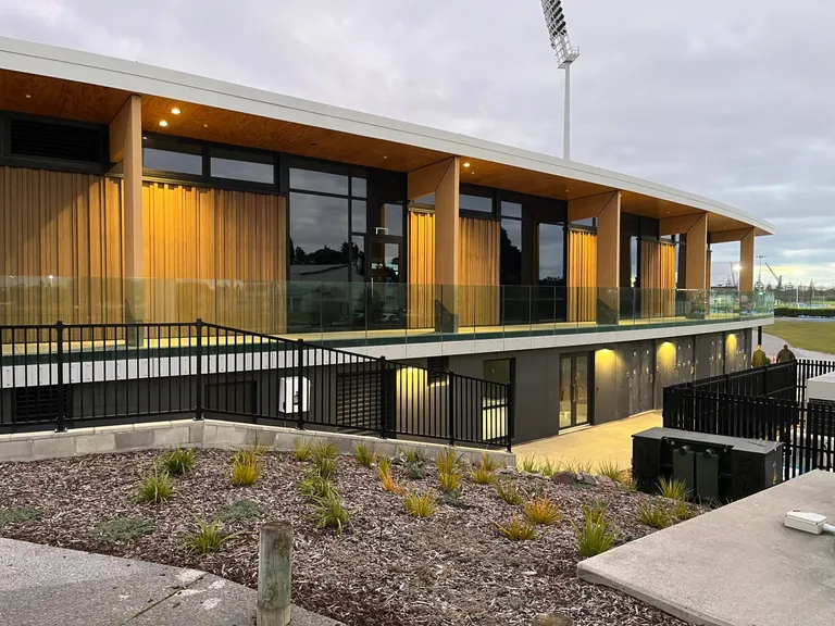 Bay Oval pavilion at dusk: a curved timber soffit over a glass-balustraded terrace, with the ground floodlight behind
