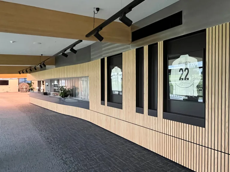 Interior of the Bay Oval pavilion: a curved servery counter beside a timber batten wall displaying framed cricket shirts